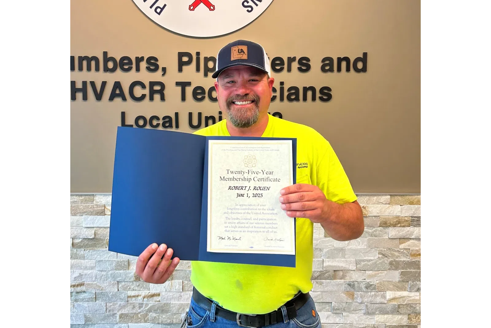 Smiling man in yellow shirt holding a certificate standing beside a Plumbers and Pipefitters union sign on wall.