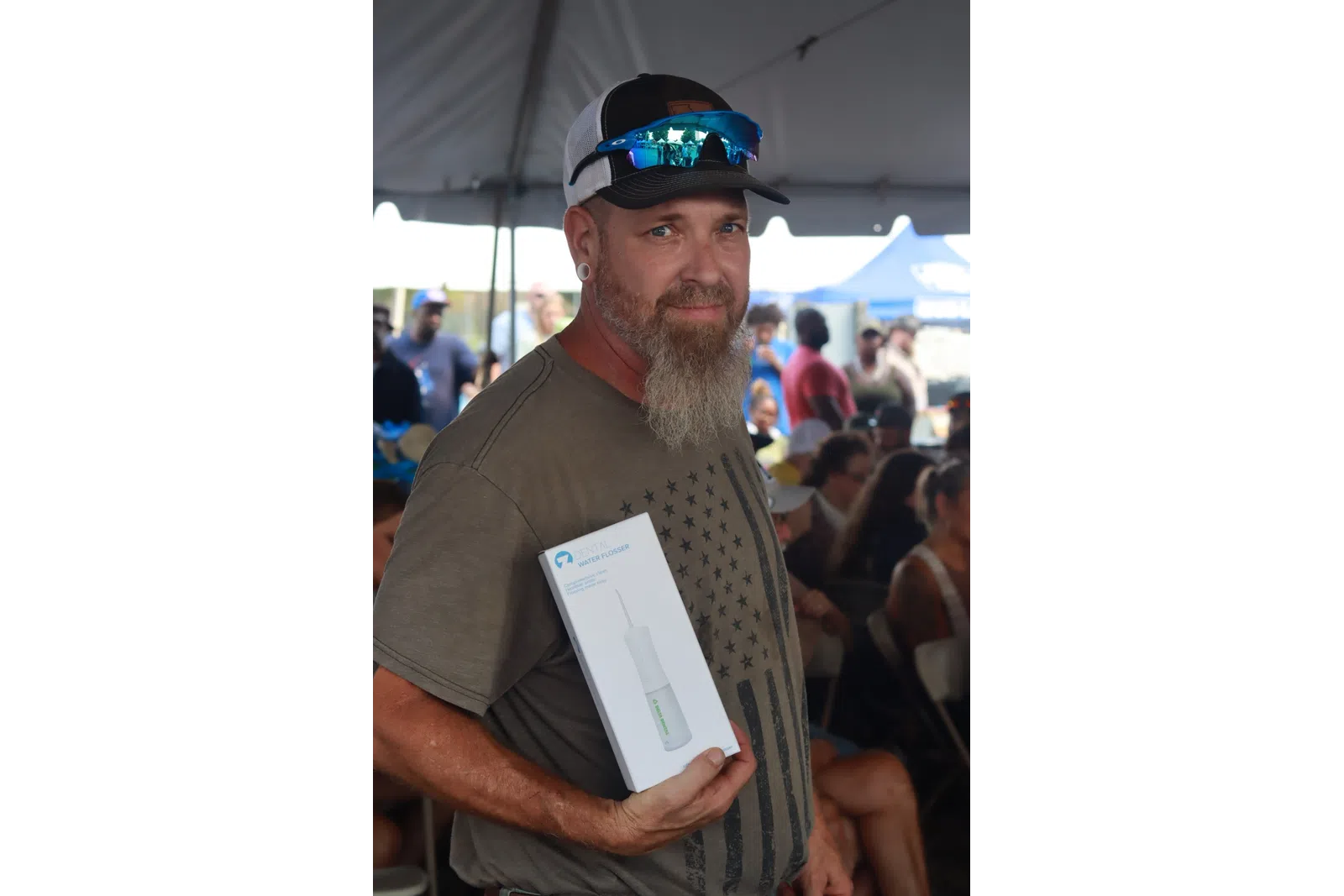 Man with a beard and sunglasses holding a white product box in a crowded event tent