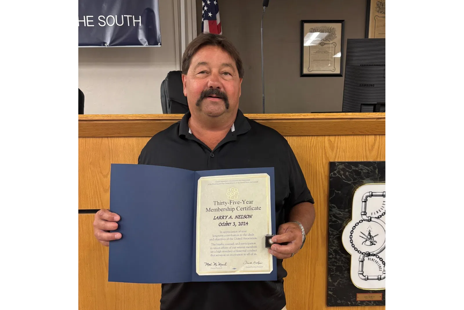 Man in black shirt and shorts holding a certificate in a formal office with wooden paneling and an American flag.