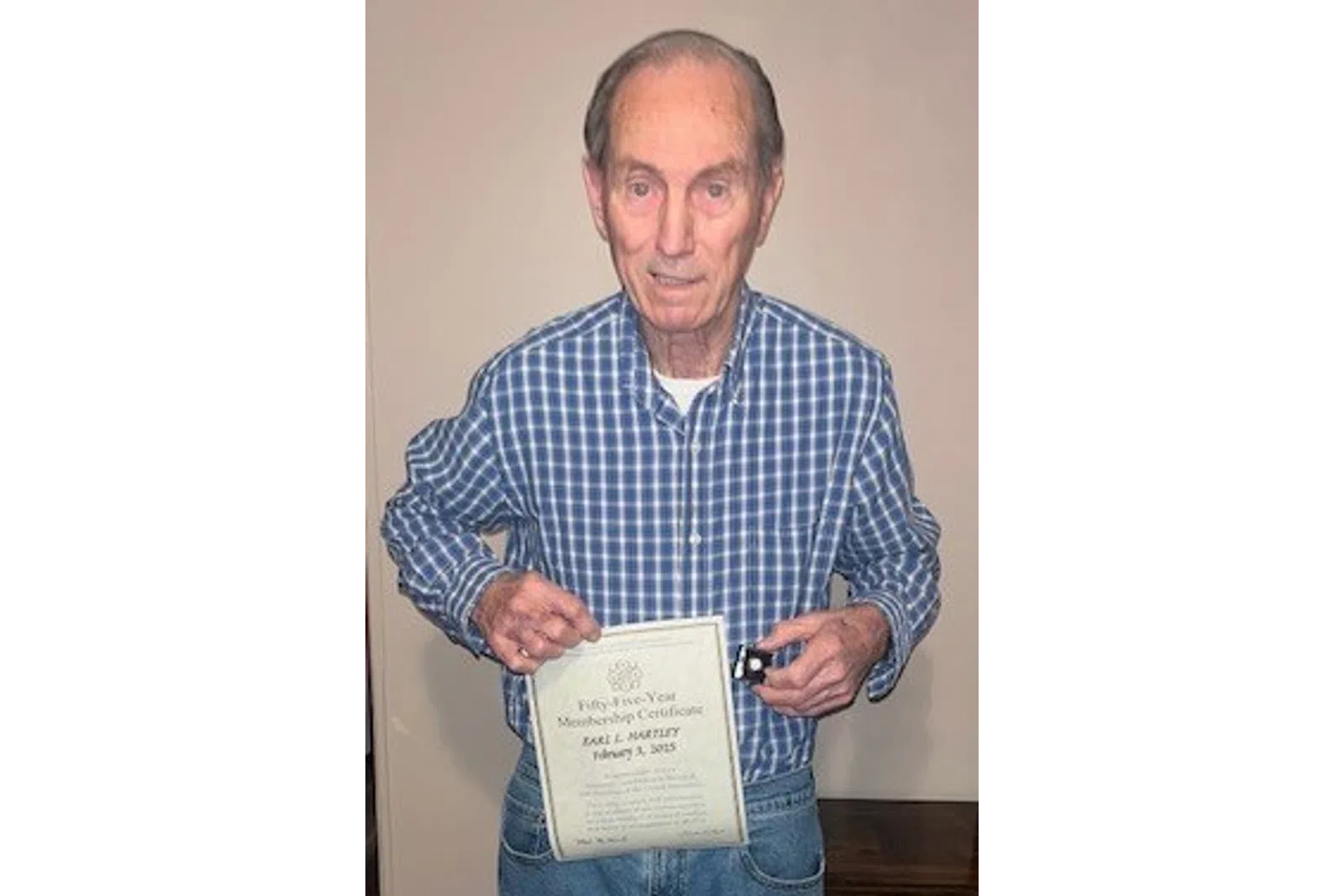 Elderly man in blue plaid shirt holding a certificate and a small black box, standing indoors on wood floor.