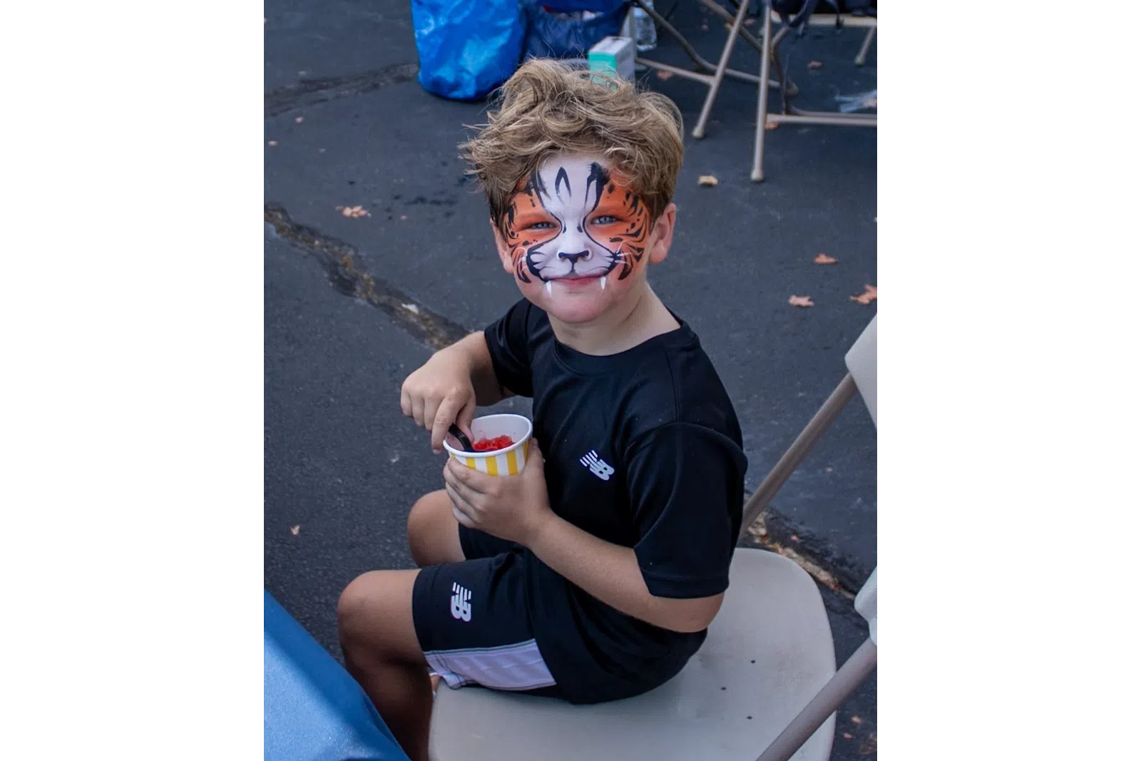 Young boy with tiger face paint sitting on a chair holding a cup of snacks outdoors at a community event.