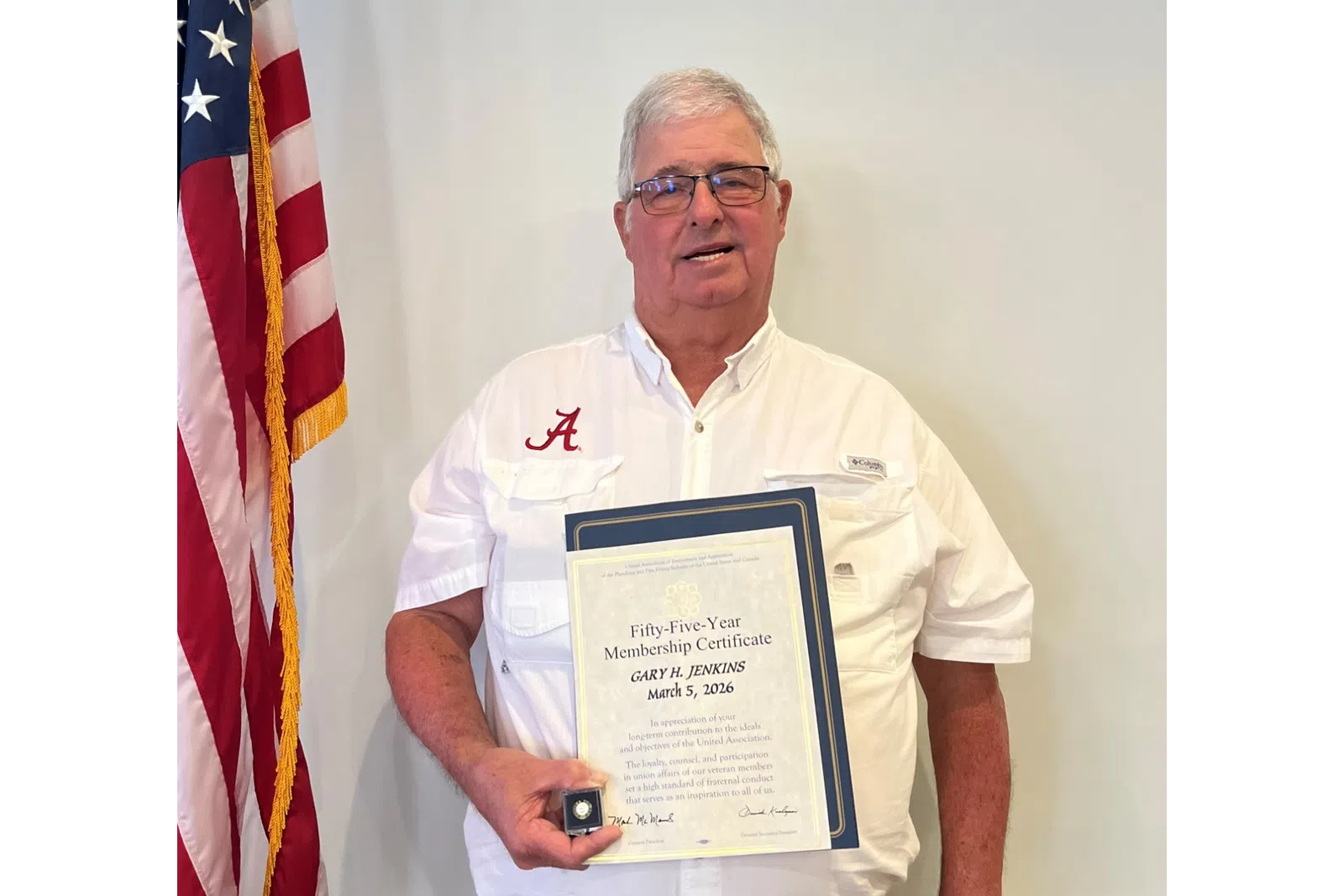 Man in white shirt holding a certificate and medal standing next to an American flag indoors.
