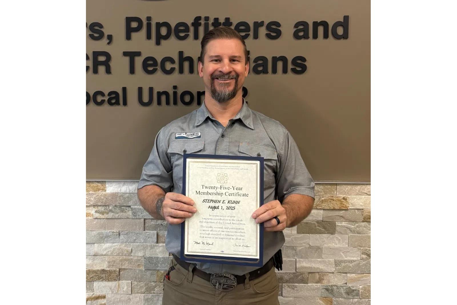 Man in uniform holding certificate standing by Plumbers and Pipefitters Local Union 72 sign on wall.