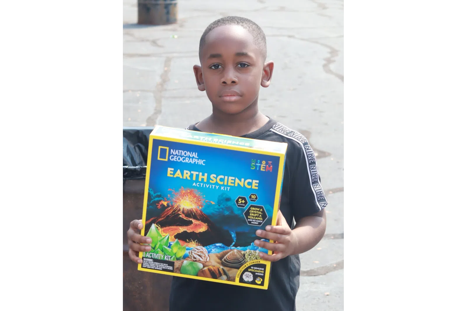 Young boy holding a National Geographic Earth Science activity kit standing outdoors on a paved area.