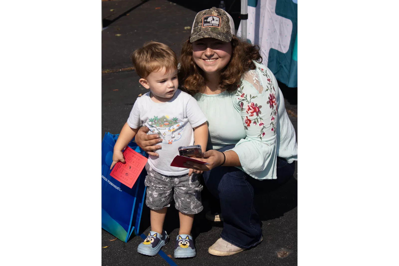 Smiling woman with floral blouse and camo hat kneels next to toddler holding red paper and phone outside on pavement.