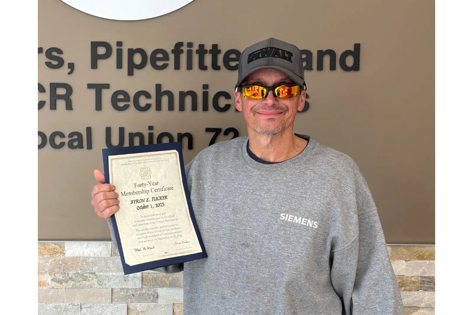 Man wearing sunglasses and a Siemens sweatshirt holding a certificate next to a Plumbing and HVAC union sign.
