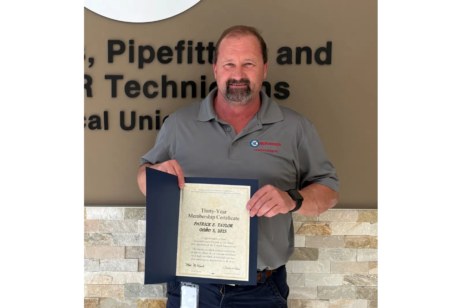 Man proudly holding certificate standing by Plumbers, Pipefitters & Service Technicians union sign on wall