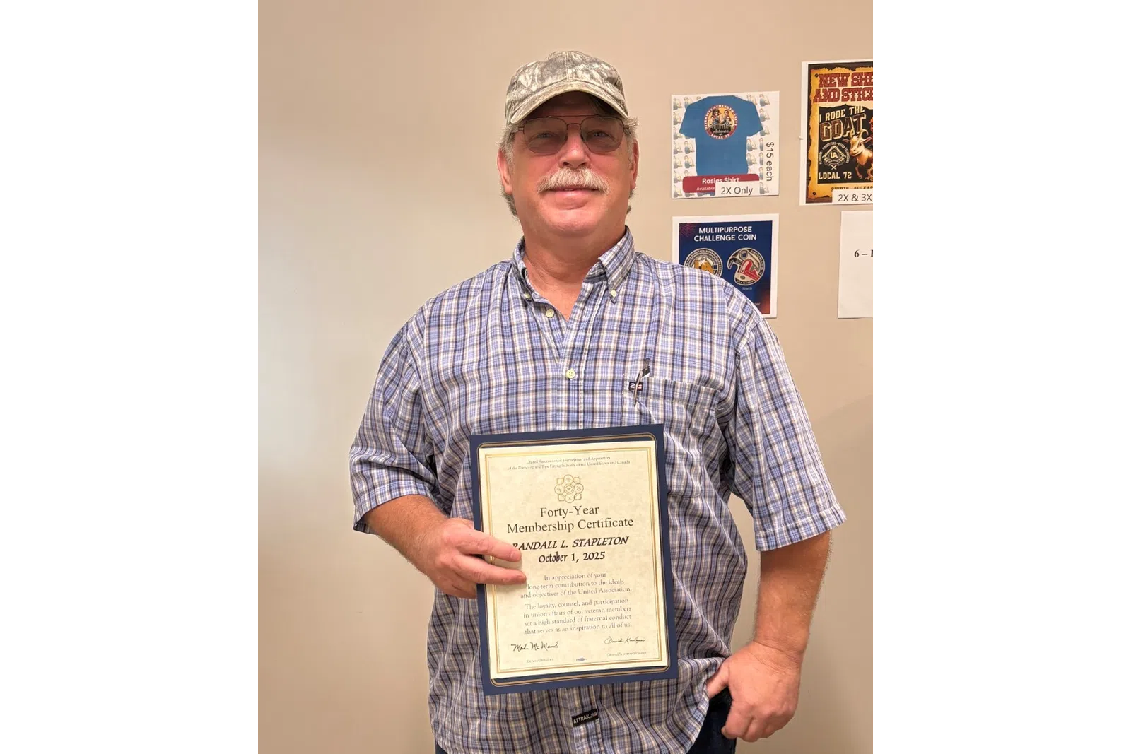 Man in plaid shirt and cap holding a framed certificate standing in a room with posters on the wall.
