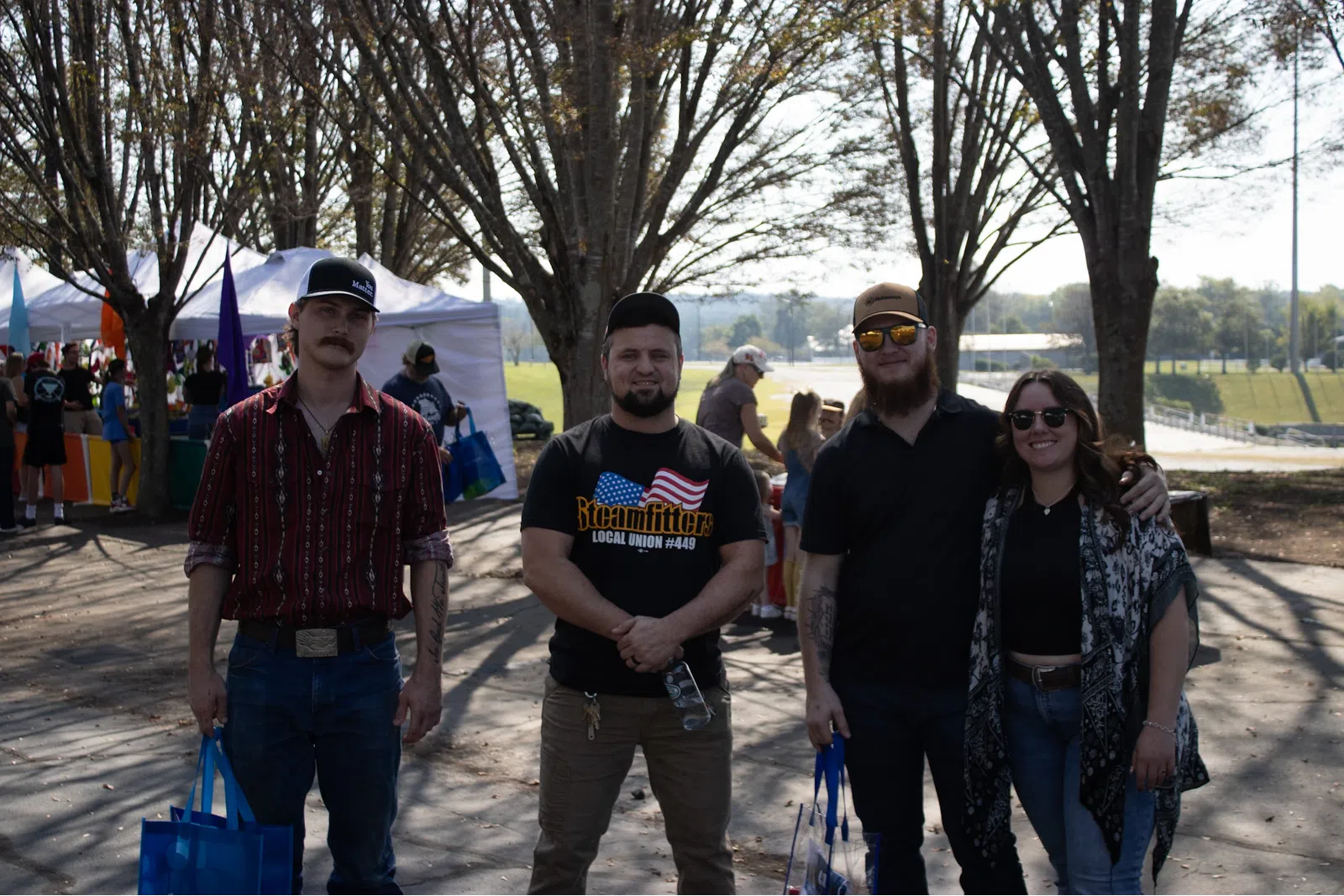 Four friends standing outdoors at a sunny event with tents and trees in the background, holding blue bags.