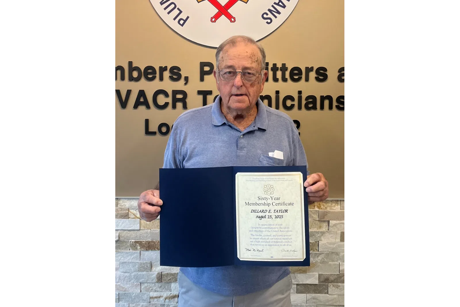 Elderly man holding a certificate standing next to a Plumbers and Pipefitters union emblem on a beige wall.