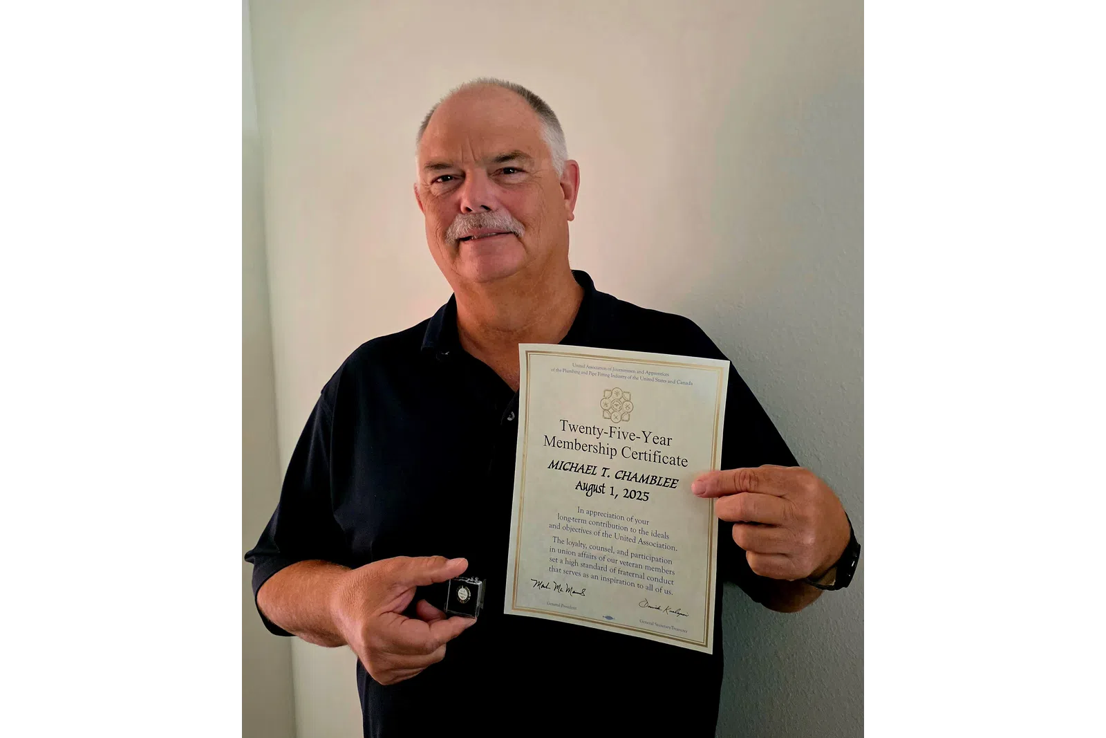 Man holding a Twenty-Five-Year Membership Certificate and a lapel pin in front of a plain wall.