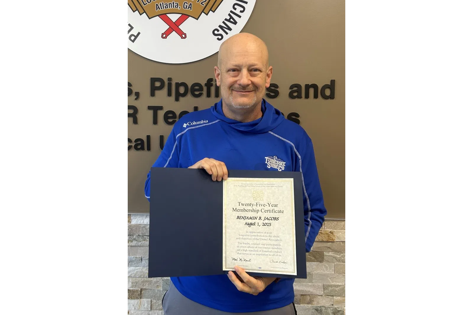 Man in blue jacket holding certificate in front of Plumbers and Pipefitters Union Local 72 logo and wall text