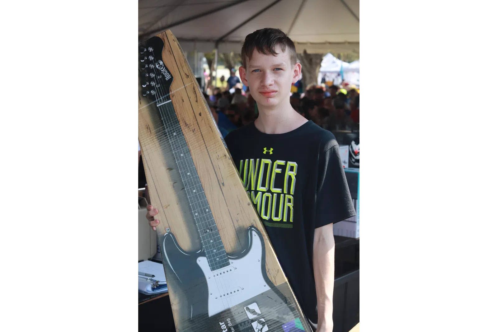 Boy holding boxed electric guitar at an outdoor event with tent and crowd in the background
