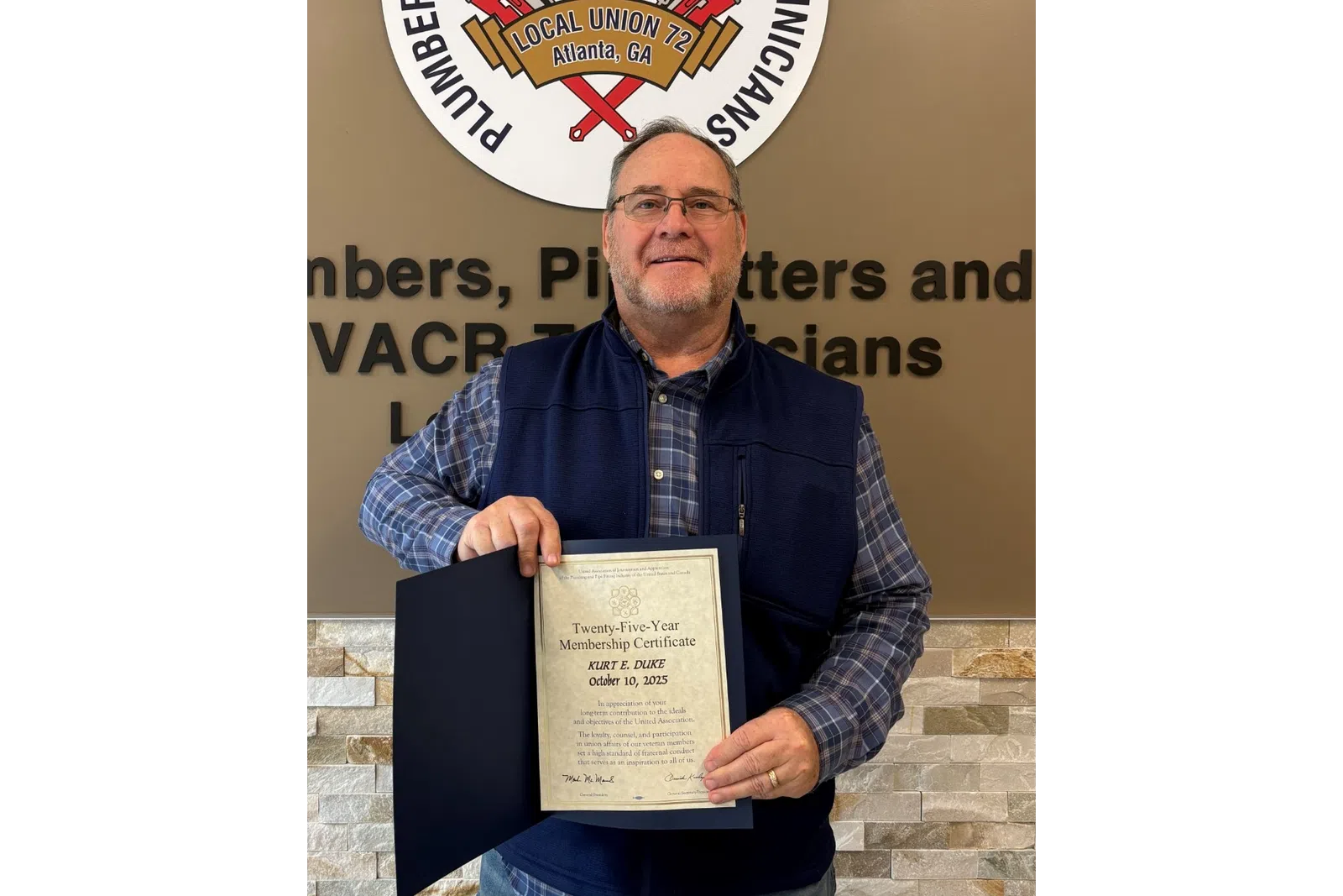 Man holding a certificate in front of a plumbers and pipefitters union sign and wall with text.