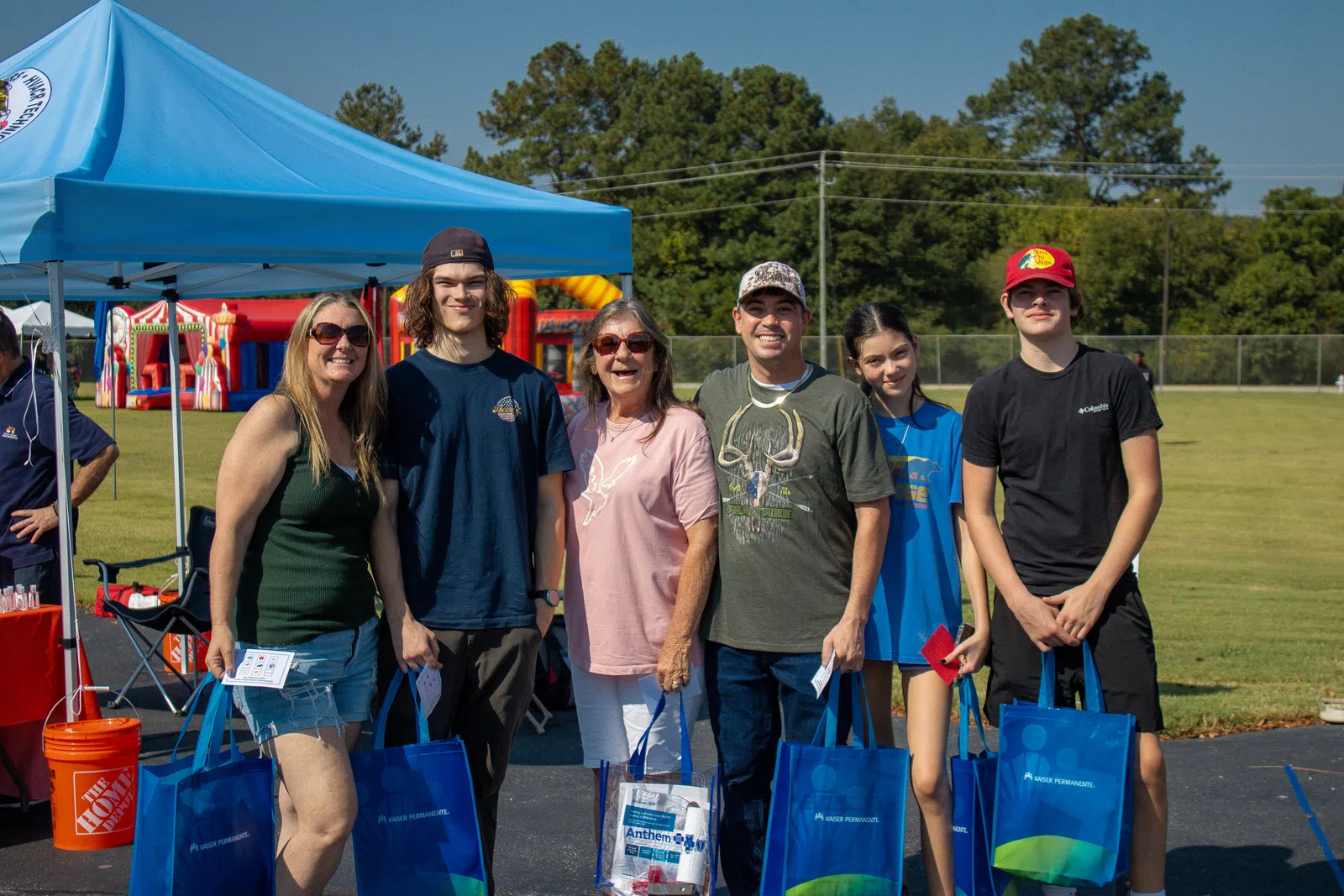 Group of six people smiling outdoors at a community event with blue tents and inflatable play structures.