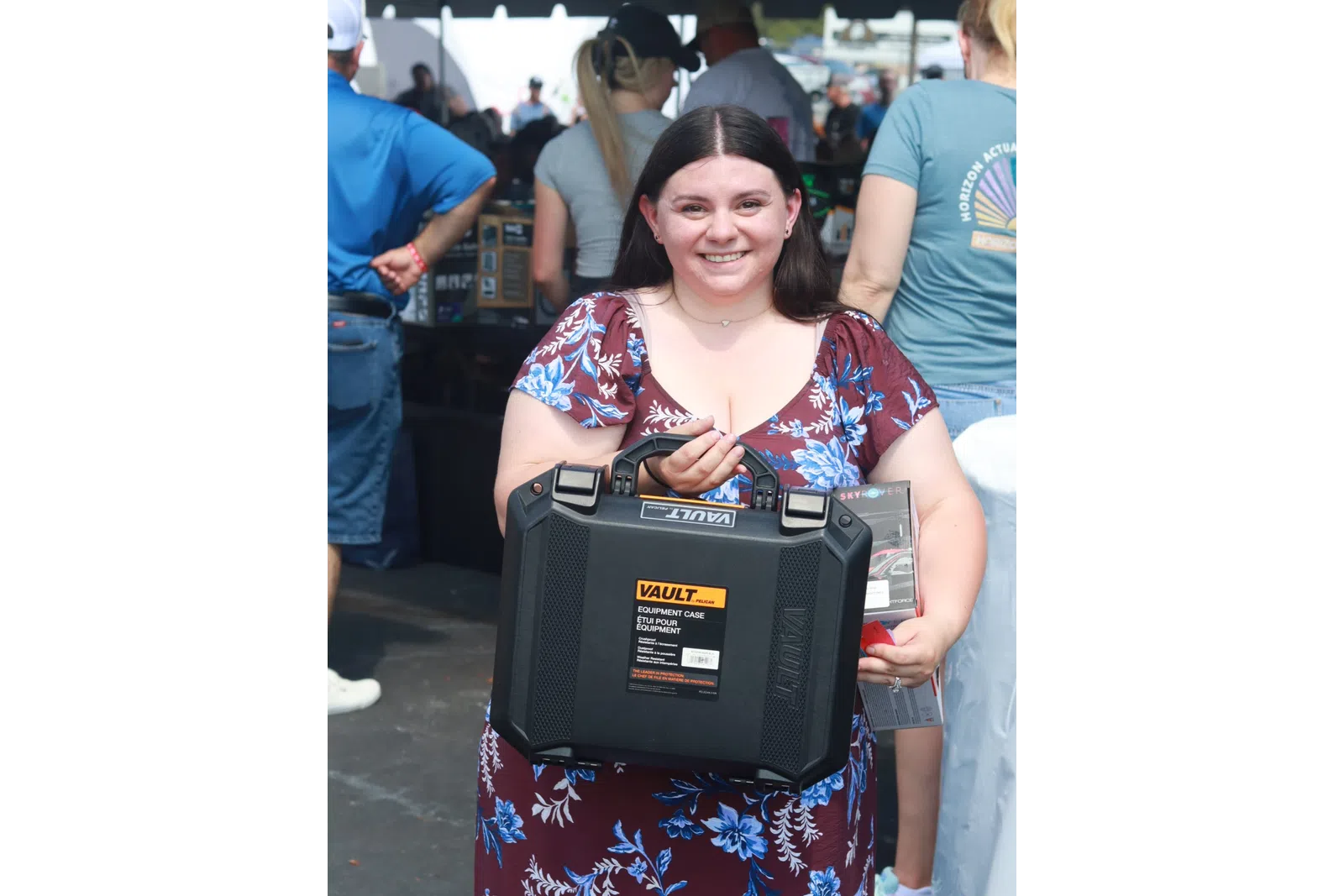 Smiling woman in floral dress holding a black Vault equipment case at an outdoor event.
