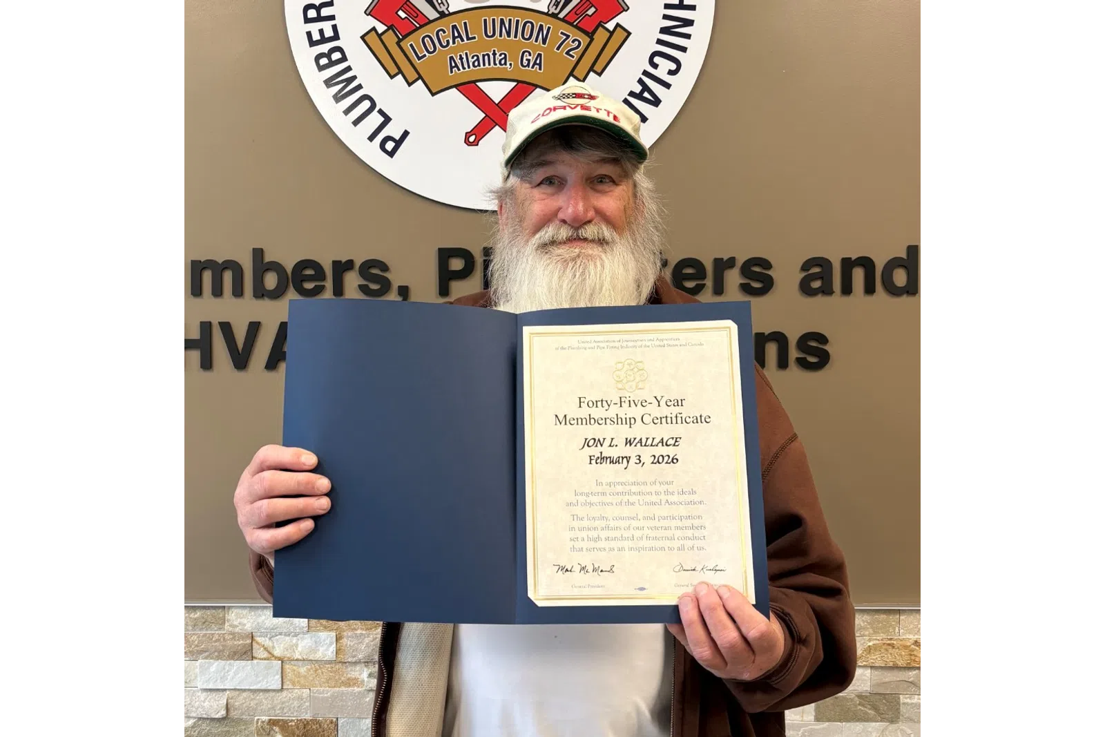 Bearded man holding a certificate in front of Plumbers and Pipefitters Local Union 72 sign on wall.