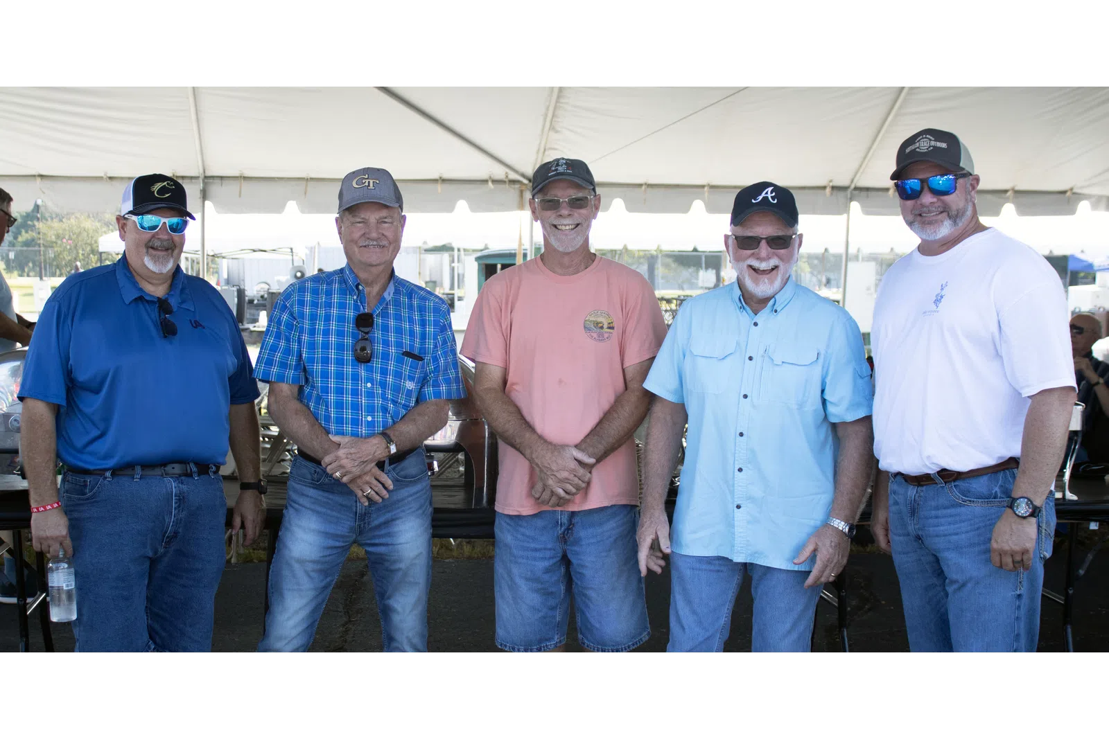Five men wearing casual clothes and caps smiling under a white event tent on a sunny day.