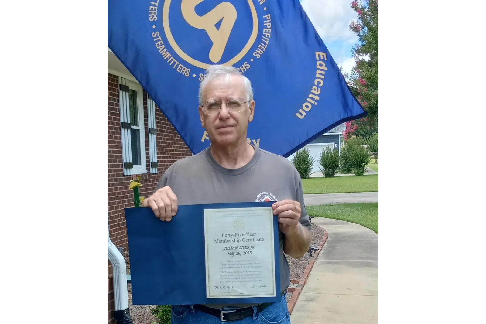 Man holding a Forty-Five-Year Membership Certificate outdoors with a blue flag in the background