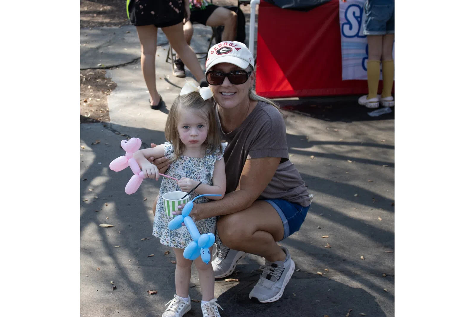 Young girl holding pink and blue balloon animals with woman in sunglasses and cap crouching beside her outdoors.