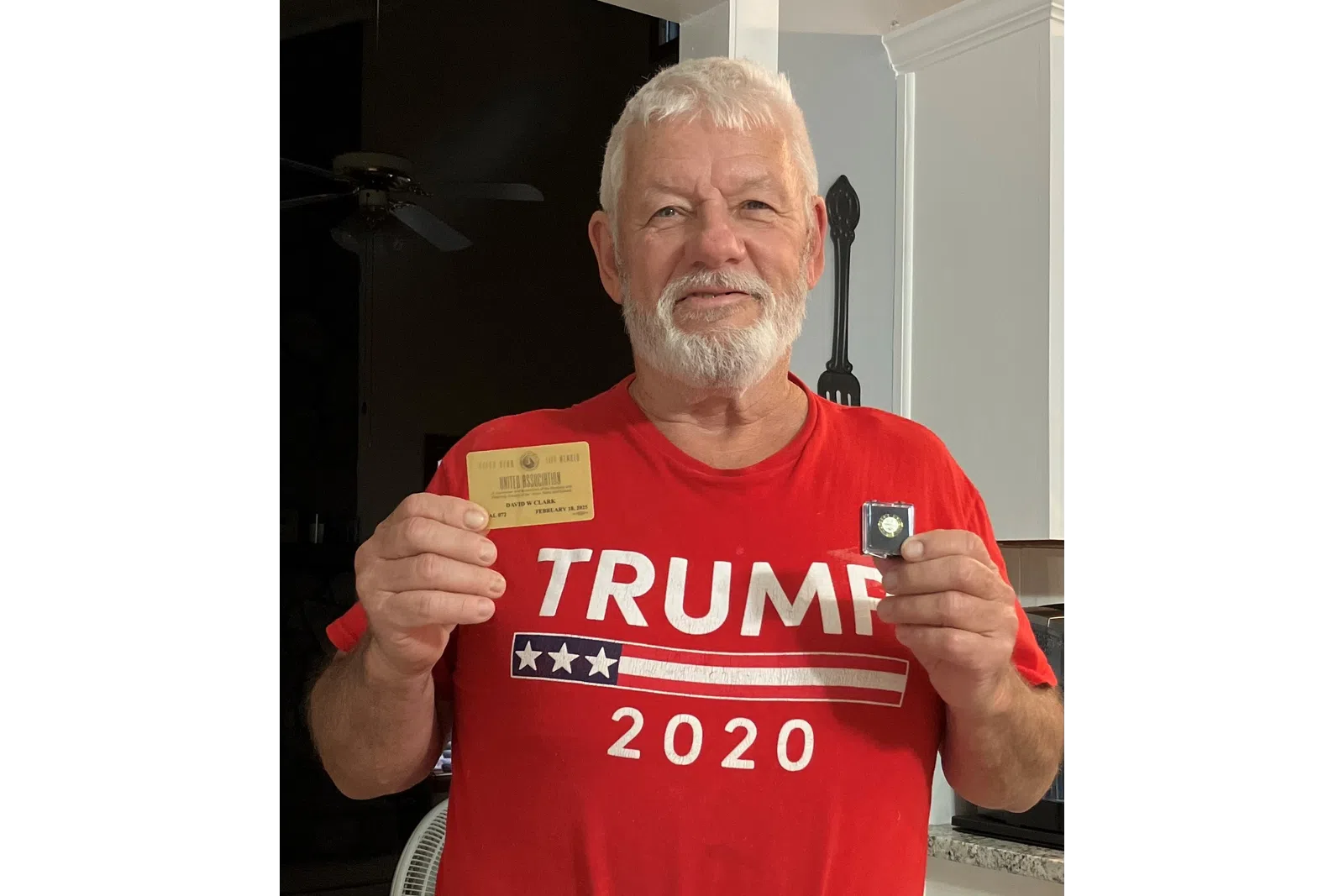 Older man with white beard wearing a red Trump 2020 shirt holding a card and a small medal in a kitchen.