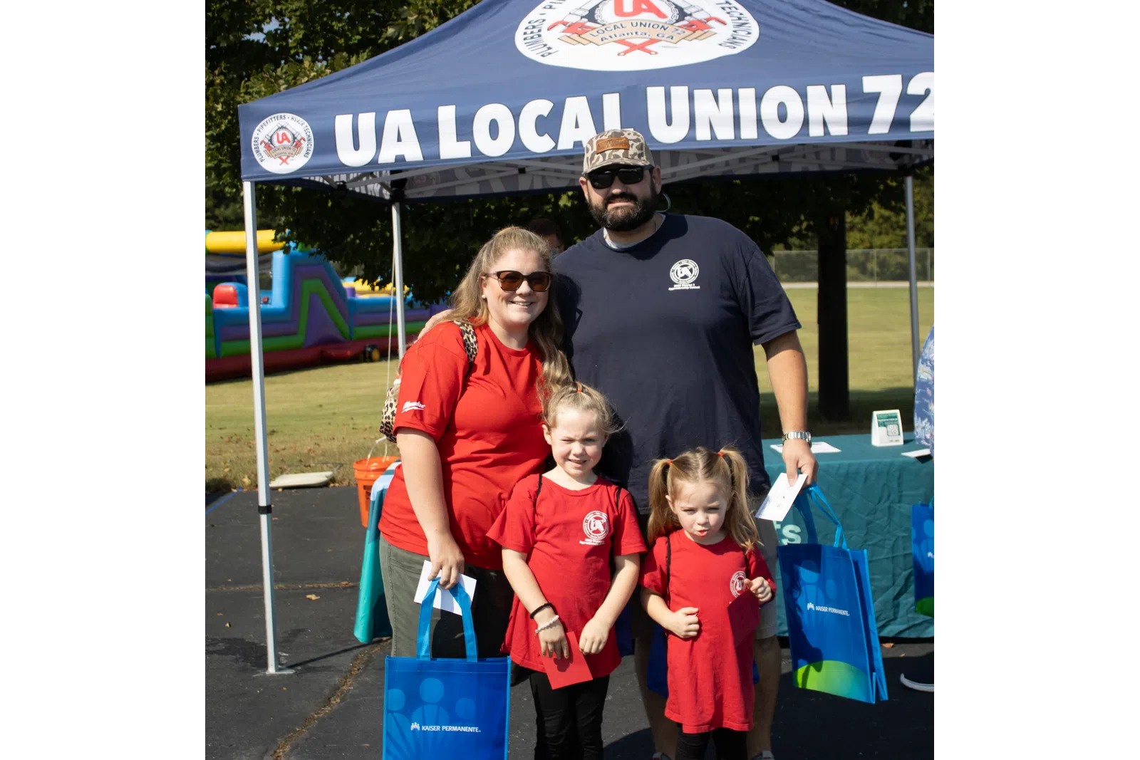 Family of four standing under UA Local Union 72 tent holding blue Kaiser Permanente bags outdoors.