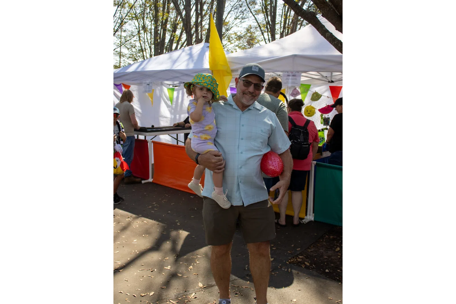 Smiling man holding a toddler in a colorful hat at an outdoor market with tents and festive decorations.