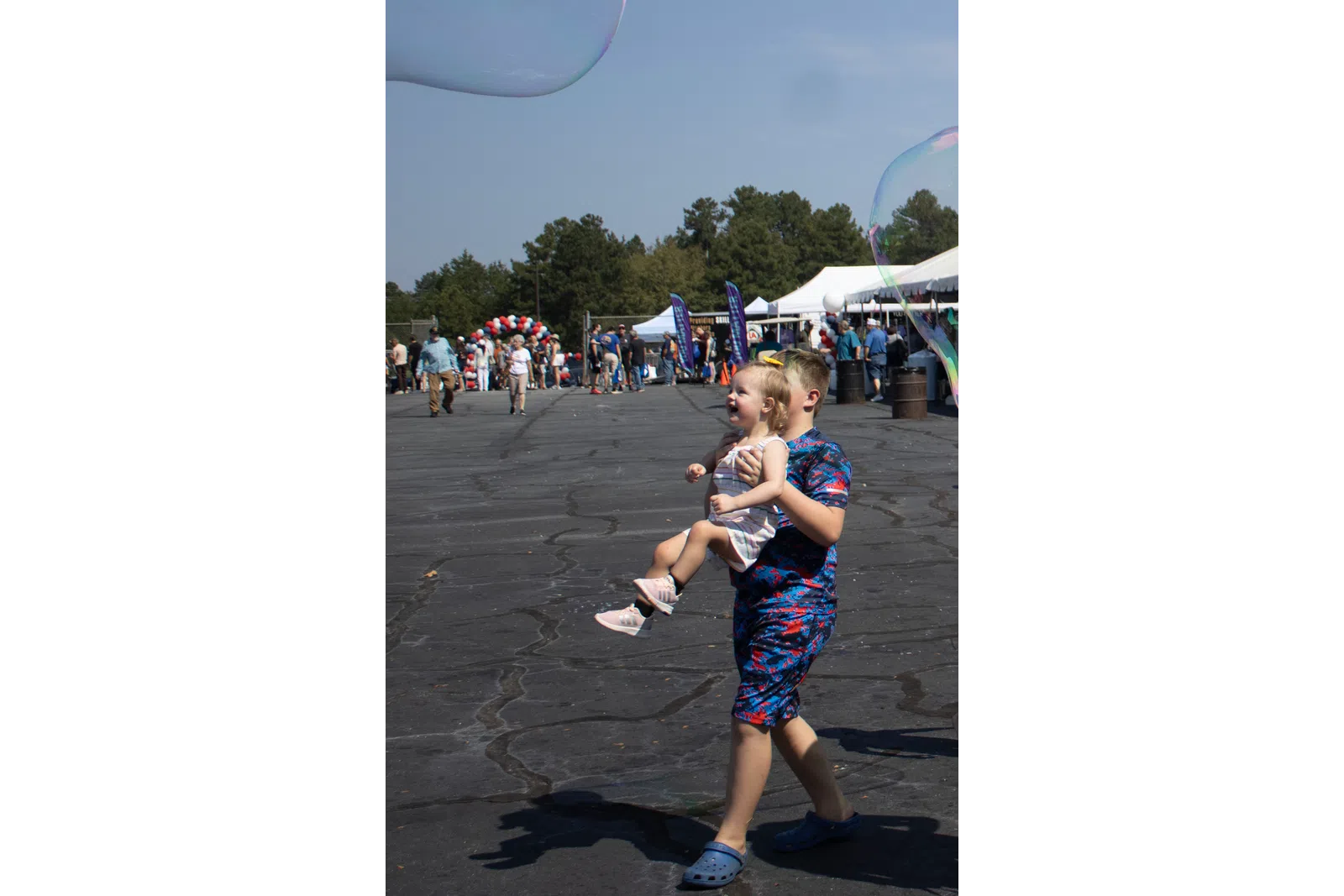 Boy carrying a smiling toddler girl outdoors with tents and people in the background on a sunny day.