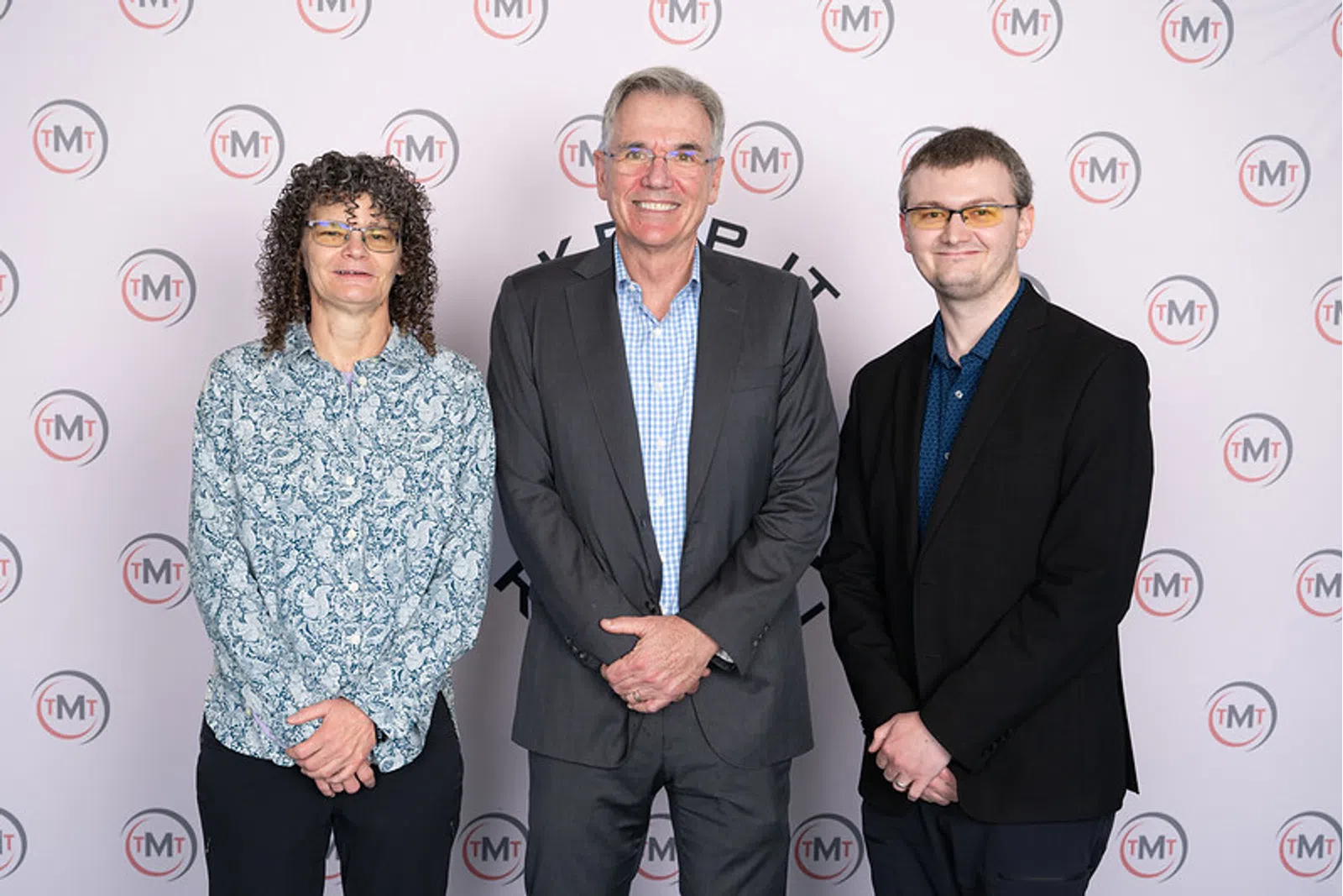 Three people posing professionally in front of a backdrop with TMT logos, dressed in business and casual attire.