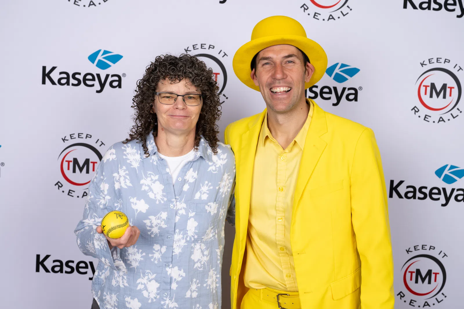 Two people posing at a Kaseya event, one in a yellow suit and hat holding a yellow softball, the other in a floral shirt.