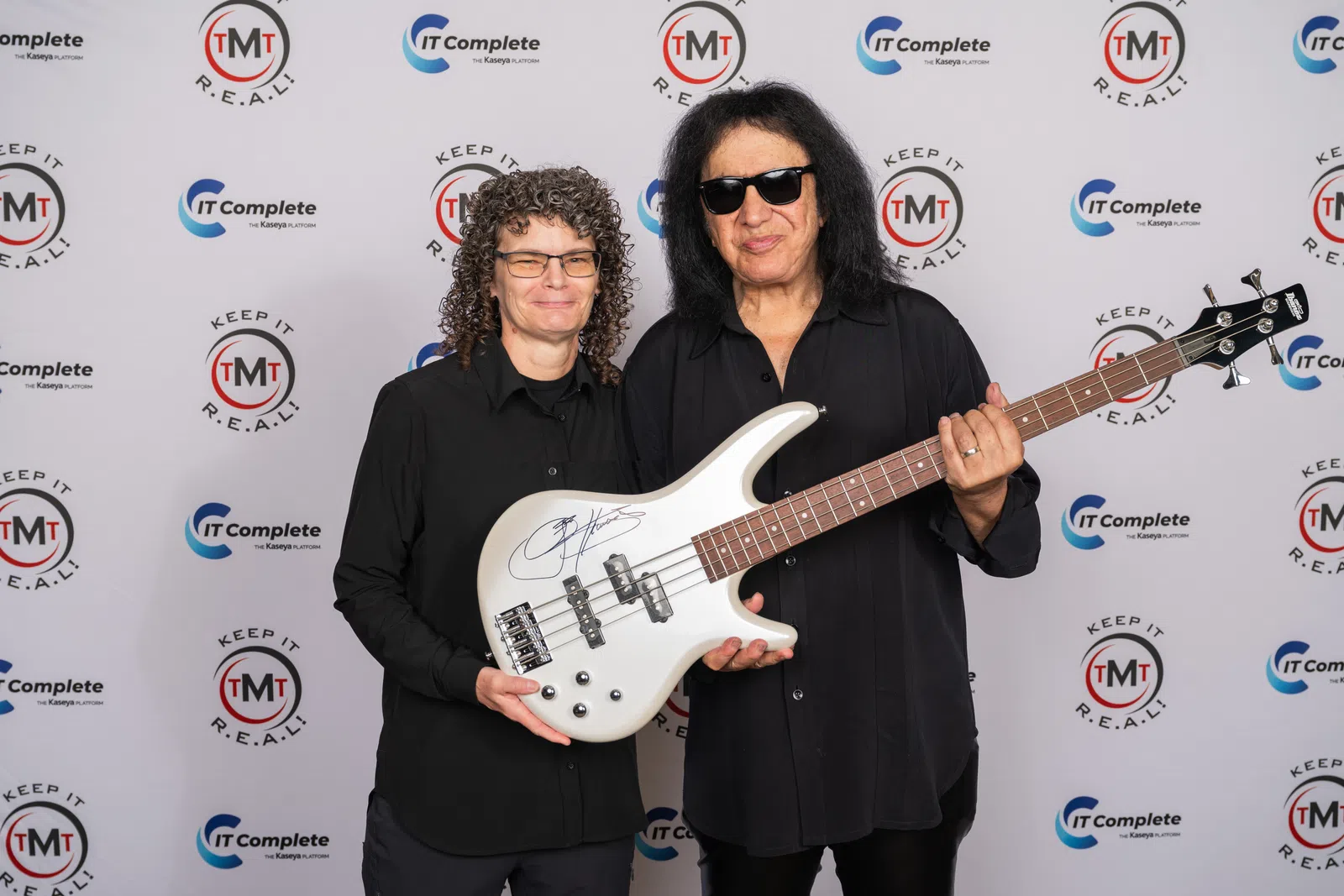 Two people holding a signed white electric bass guitar in front of a branded event backdrop.