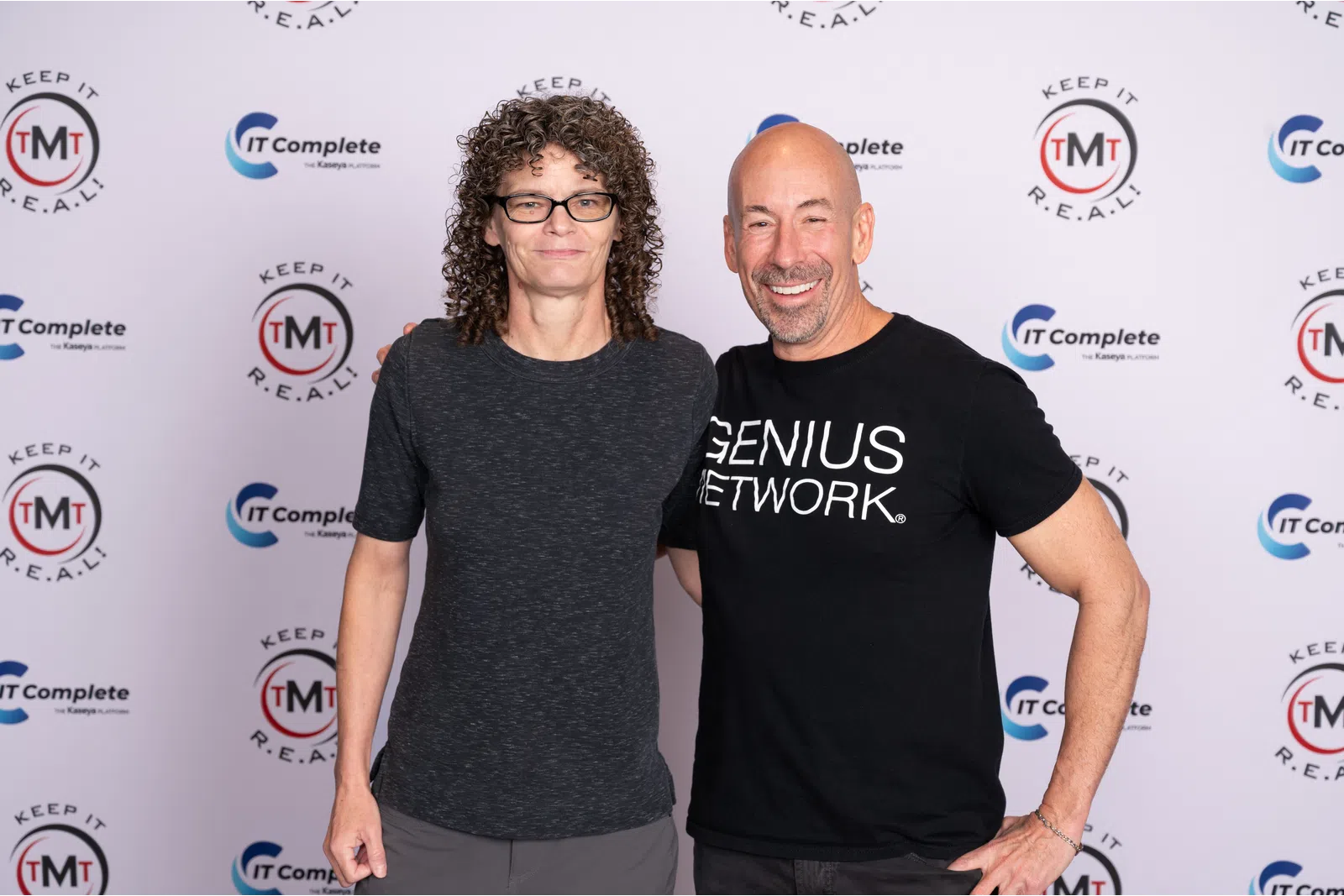 Two smiling adults posing in front of a branded backdrop with logos and slogans for IT Complete and TMT Keep It Real