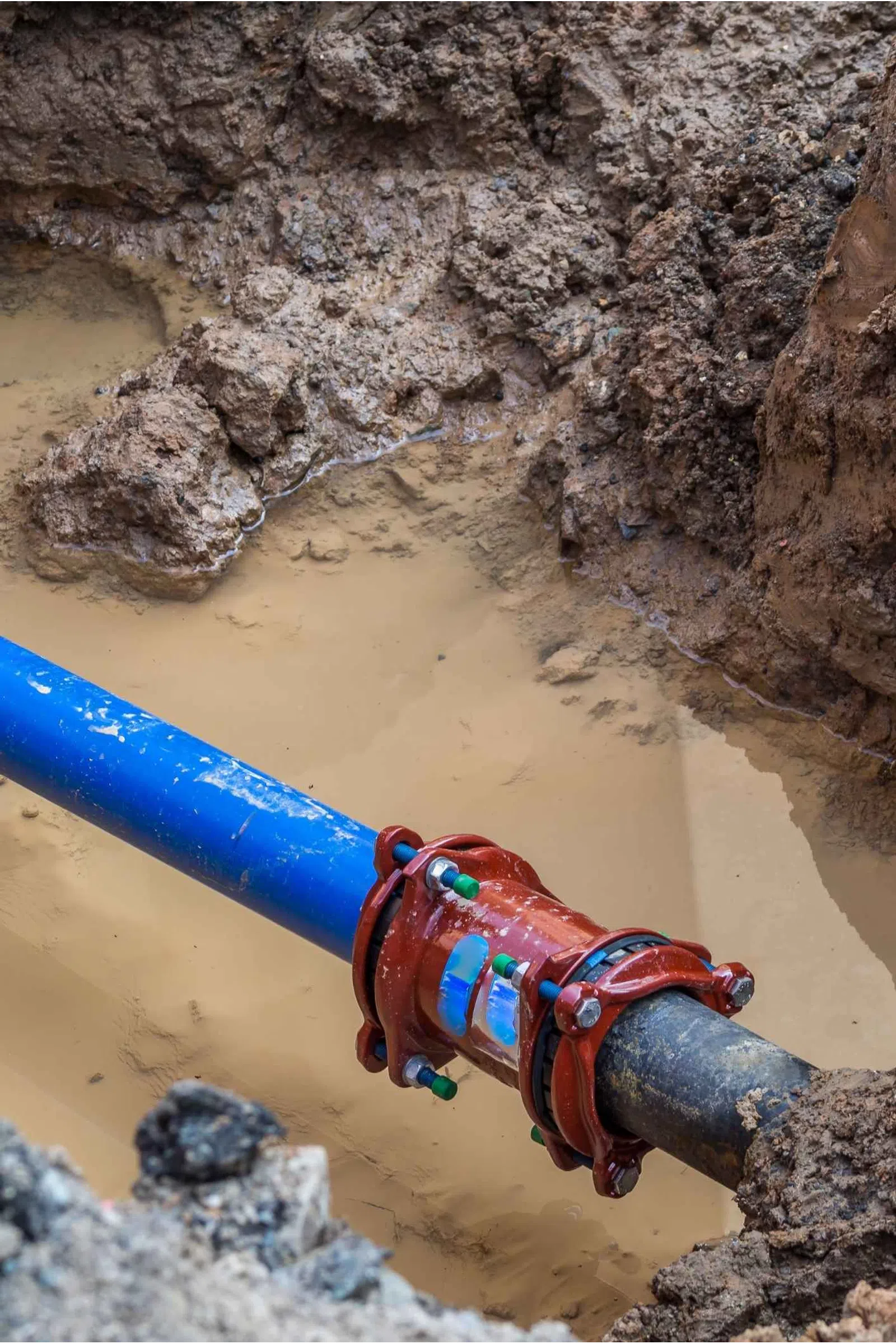 Blue and black pipes connected by a red joint fitting in a muddy trench with water accumulation.