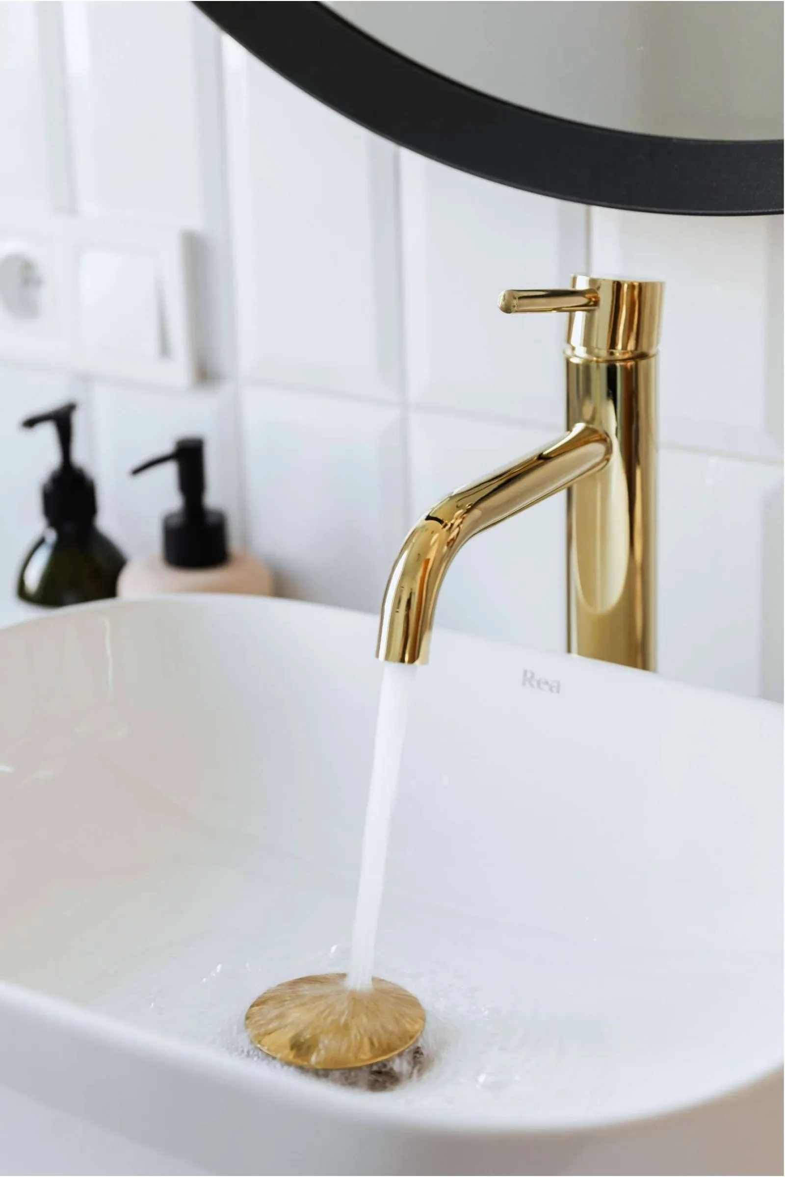 Gold faucet pouring water into a modern white bathroom sink with soap dispensers and white tiles.