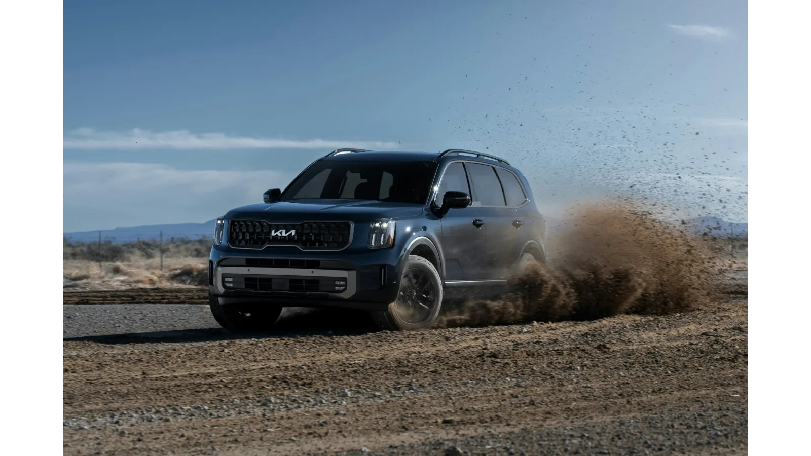 Dark blue Kia SUV kicking up dirt while driving on a dusty off-road path under a clear sky.