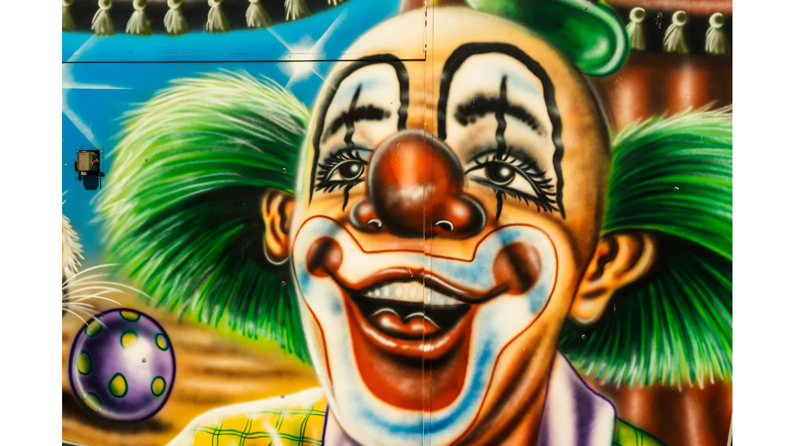 Vibrant clown mural with green hair, big red nose, and colorful makeup smiling widely on a blue background.