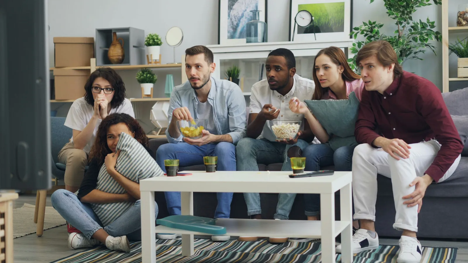 Group of diverse young adults watching TV intently while eating popcorn and snacks in a cozy living room.