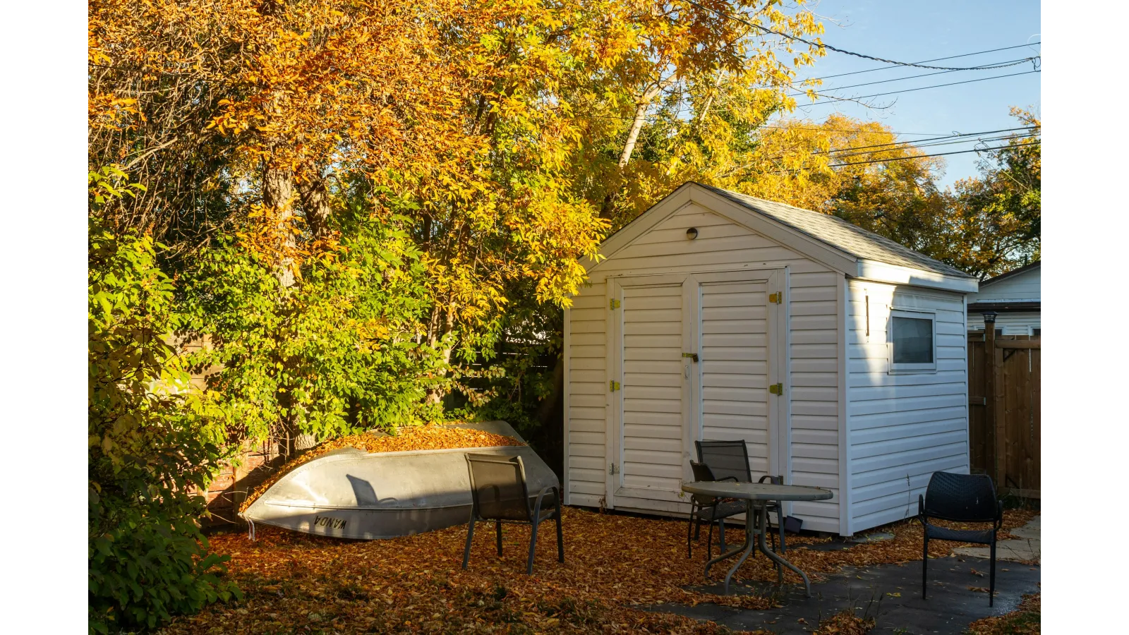White garden shed surrounded by autumn leaves, with overturned boat and outdoor chairs in backyard.