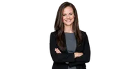 Confident young businesswoman with long brown hair in gray dress and black blazer smiling with arms crossed