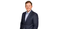 Smiling man with curly hair wearing a blue checkered blazer and light blue shirt against a white background
