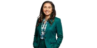 Smiling woman with long dark hair wearing a teal suit and plaid shirt posing confidently with hands in pockets.