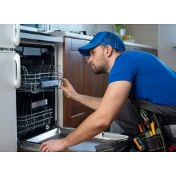 Technician in blue uniform repairing an open dishwasher in a kitchen setting with tools in a belt.