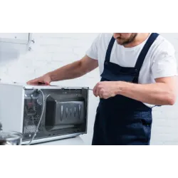 Technician repairing a microwave oven, wearing blue overalls and working on the appliance's interior components.