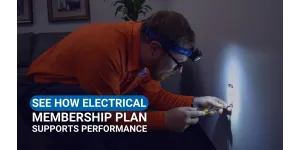 Electrician in orange shirt fixing wall outlet with headlamp light focused on work area indoors.