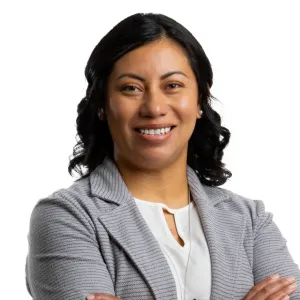 Confident woman with dark curly hair smiling, wearing a gray blazer and white blouse, arms crossed.