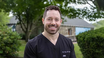 Smiling man in black medical scrubs standing outdoors with trees and a stone building in the background