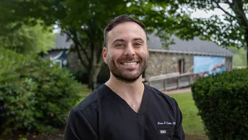 Smiling man in a black scrub top stands outdoors by greenery and a stone building on a bright day.