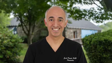 Smiling middle-aged male doctor in black scrubs standing outdoors with trees and a building in the background.
