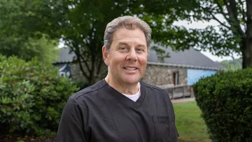 Middle-aged man in black scrub top smiling outdoors with trees and a stone building in the background