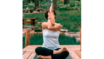 Woman practicing eagle pose yoga on a wooden deck surrounded by green nature and serene outdoor setting